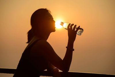 Side view of silhouette woman drinking water from bottle against orange sunset sky