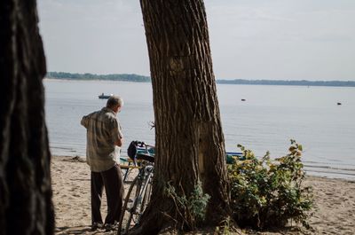 Rear view of man standing by tree trunk at beach against sky