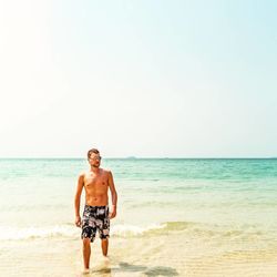 Full length of shirtless man walking at beach against sky in sunny day