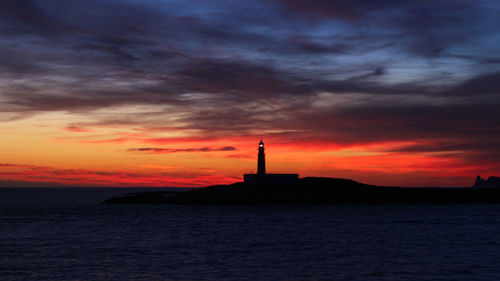 Silhouette lighthouse by sea against orange sky