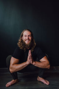 Smiling handsome flexible male athlete in sportswear with namaste performing yoga kakasana position on wooden floor against black wall of contemporary workout room