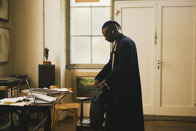 Side view of businessman in overcoat packing backpack while standing in workshop