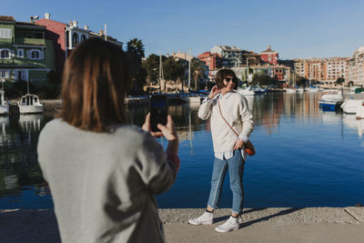 Rear view of woman photographing smiling friend standing by river in city