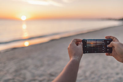 Midsection of person holding camera at beach against sky during sunset