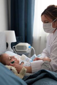 Female doctor wearing mask examining girl sleeping on bed