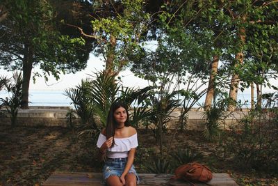 Portrait of smiling young woman sitting on bench against plants at park