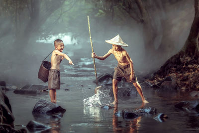 Shirtless siblings fishing at river in forest