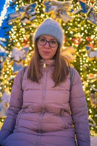 Portrait of young woman standing against illuminated christmas tree