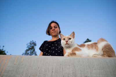 Low angle view of teenager looking at stray cat sitting on retaining wall against sky