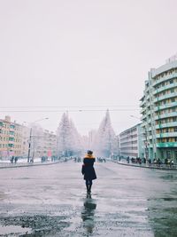 Full length rear view of woman standing on wet city street against clear sky during winter