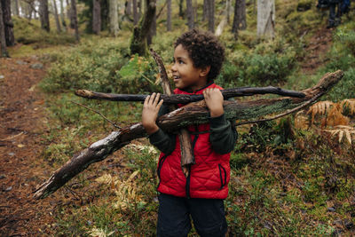 Smiling boy looking away while carrying firewood in forest