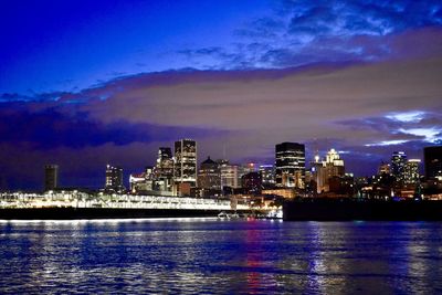 Illuminated buildings by river against sky at dusk