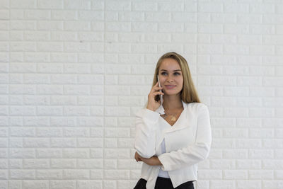Young businesswoman talking on phone while standing against white brick wall