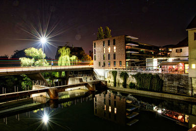 Illuminated bridge over river by buildings against sky at night