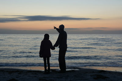 Silhouette father with daughter standing at beach against sky during sunset