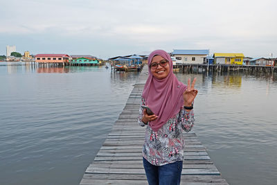 Portrait of smiling woman wearing hijab showing thumbs up while standing on pier