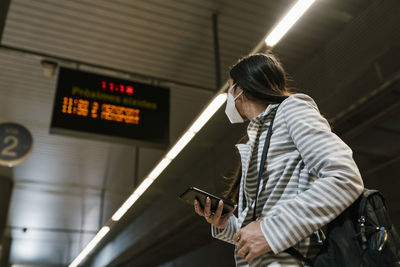 Woman looking away while standing with smart phone waiting at railroad station