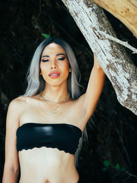 Portrait of young trans woman standing against tree trunk