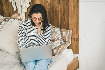 A woman sits at home on the bed talking on the phone and looking into a laptop.