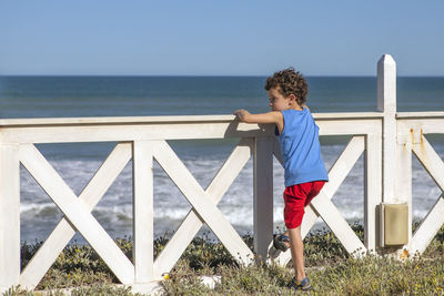 Curly-haired caucasian boy leaning on fence looking out to sea