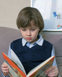 Cute boy reading book sitting on sofa at home