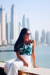 Young woman sitting by sea with modern buildings in background