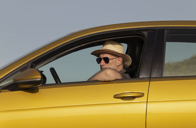 Adult man in hat and sunglasses sitting in car against clear sky