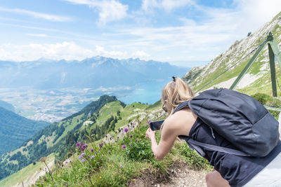 Mature woman with backpack taking a photo with smartphone of flowers on a mountain with swiss alps
