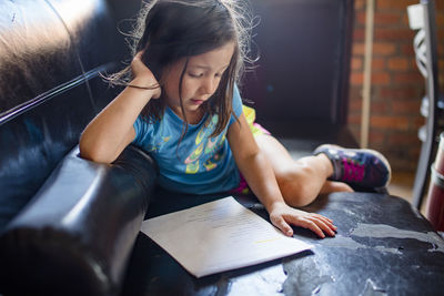 A little girl lays on a worn leather couch in sunlight reading