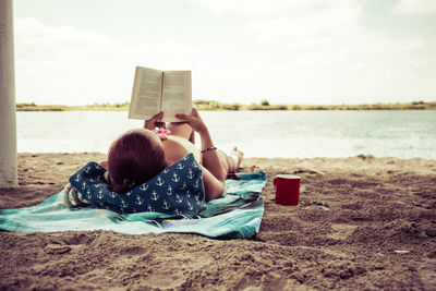 Rear view of woman reading book while relaxing on sandy beach in summer day. copy space.