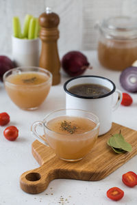 Homemade bone broth in mugs with spices on a wooden stick with bay leaf, tomatoes and onions.