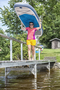Full length of happy woman carrying surfboard on pier over lake