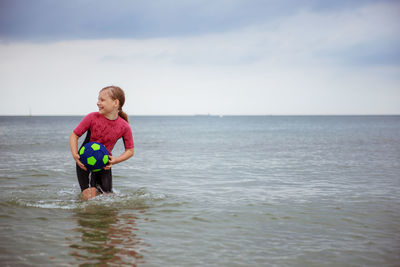 Cute girl holding soccer ball standing in sea against sky