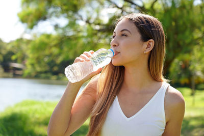 Portrait of young woman drinking water while standing against trees