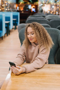 Young woman with curly hair is sitting at a cafe table and looking at her smartphone