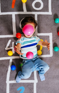 From above excited boy in casual clothes laughing and trowing up colorful balls while lying on floor with hopscotch numbers and playing at home