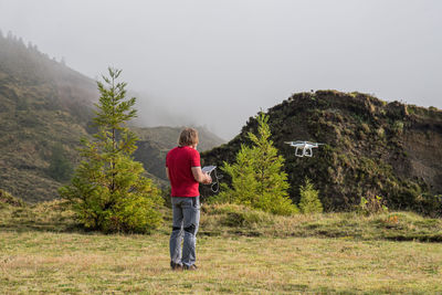 Rear view of man flying drone with remote control while standing on grass against sky