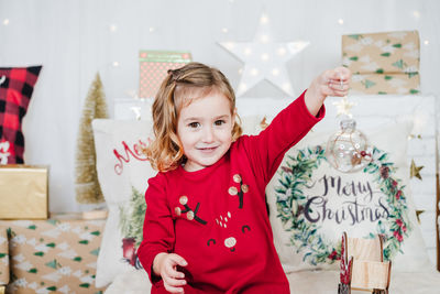 Happy little girl holding bauble wearing red christmas dress at home over christmas decoration
