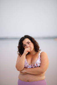 Peaceful curvy female in bikini standing on background of pink pond in summer while leaning on hand and looking at camera