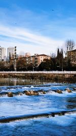 Frozen river by buildings against sky during winter