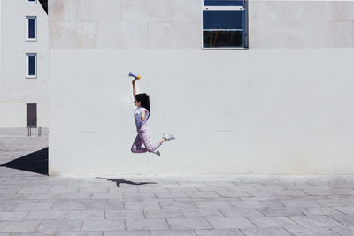 Young woman with megaphone jumping by building during sunny day