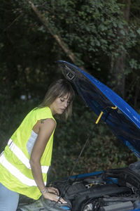 Beuty woman in reflective vest looking at engine of broken down car