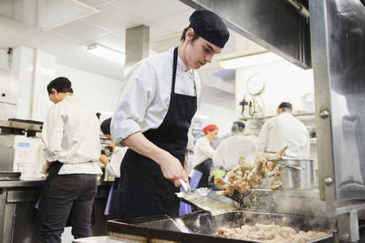 Male chef student tossing food with teacher and colleagues in background at cooking school