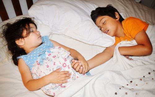 High angle view of siblings holding hands while sleeping in bed at home