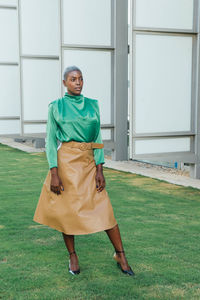 Full body of stylish confident young black lady with short hair in elegant blouse and leather skirt standing on grassy lawn in park and looking away