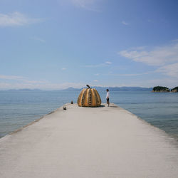 Man standing by pumpkin shaped structure on pier over sea against sky