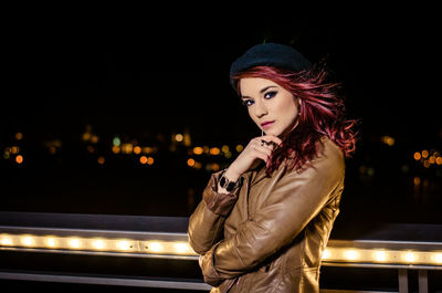 Portrait of beautiful woman standing by illuminated railing at night