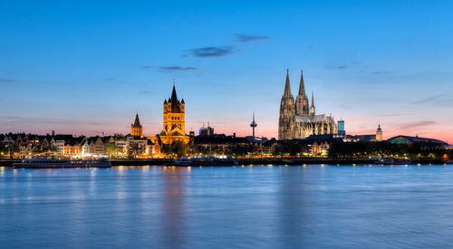 Illuminated buildings and cologne cathedral by rhine river in city at dusk