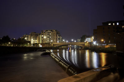 Illuminated bridge over river by buildings against sky at night