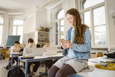 Smiling female student using smart phone while sitting at desk in classroom